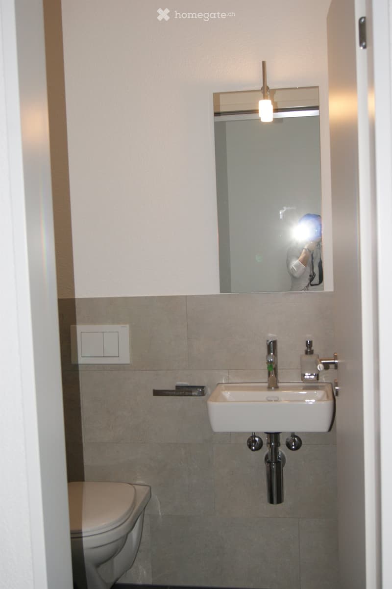Modern bathroom with a white toilet, a white sink with a chrome faucet, a mirror with a built-in light fixture, and tiled walls.