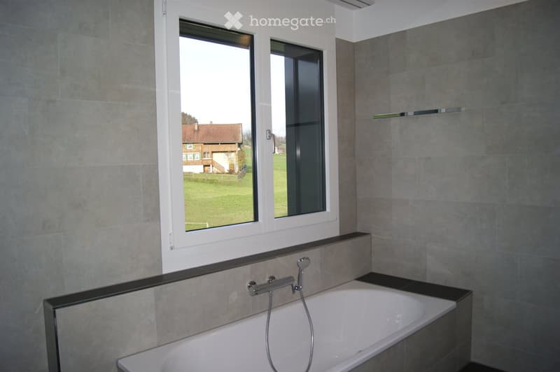 Modern bathroom with a bathtub, sleek faucet, large window offering a view of a green field and a house, grey tiled walls, and a black shelf on the right side.