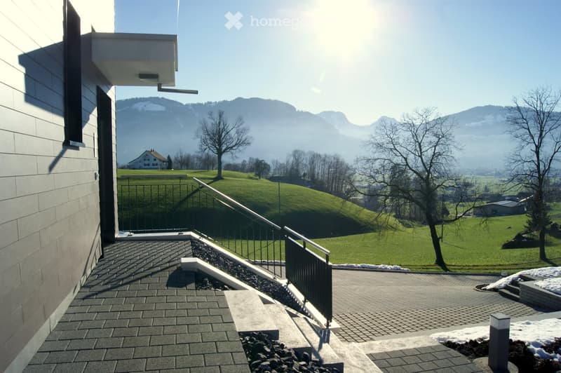 Balcony with modern railing, paved tiles, overlooking a green hillside and distant mountains.