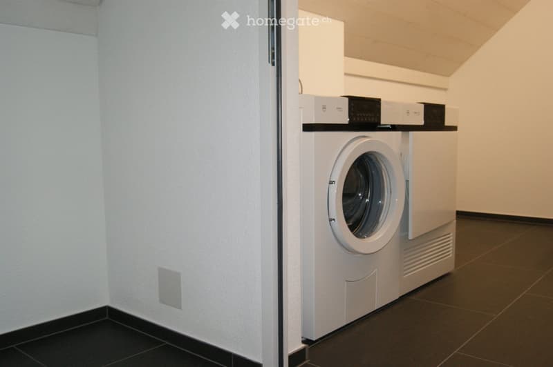 A small, modern utility room with a white washing machine and control panel mounted on the wall above it. The room features dark tiled flooring and light-colored walls.