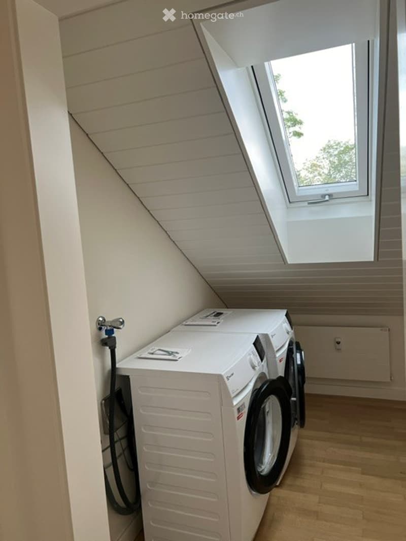A small, well-lit room with a skylight window, featuring a white washing machine and dryer unit against the wall.