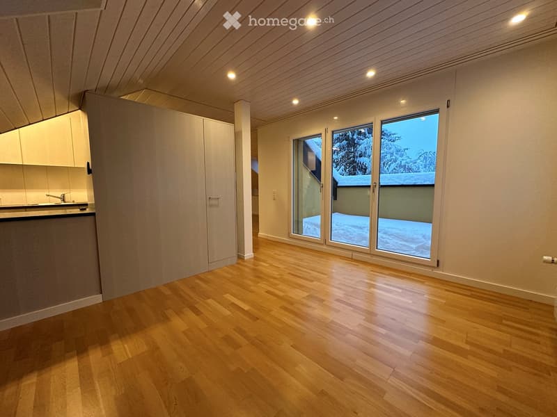Empty room with wooden floors, white walls, and large windows. There is a kitchen area with a sink and cabinets.