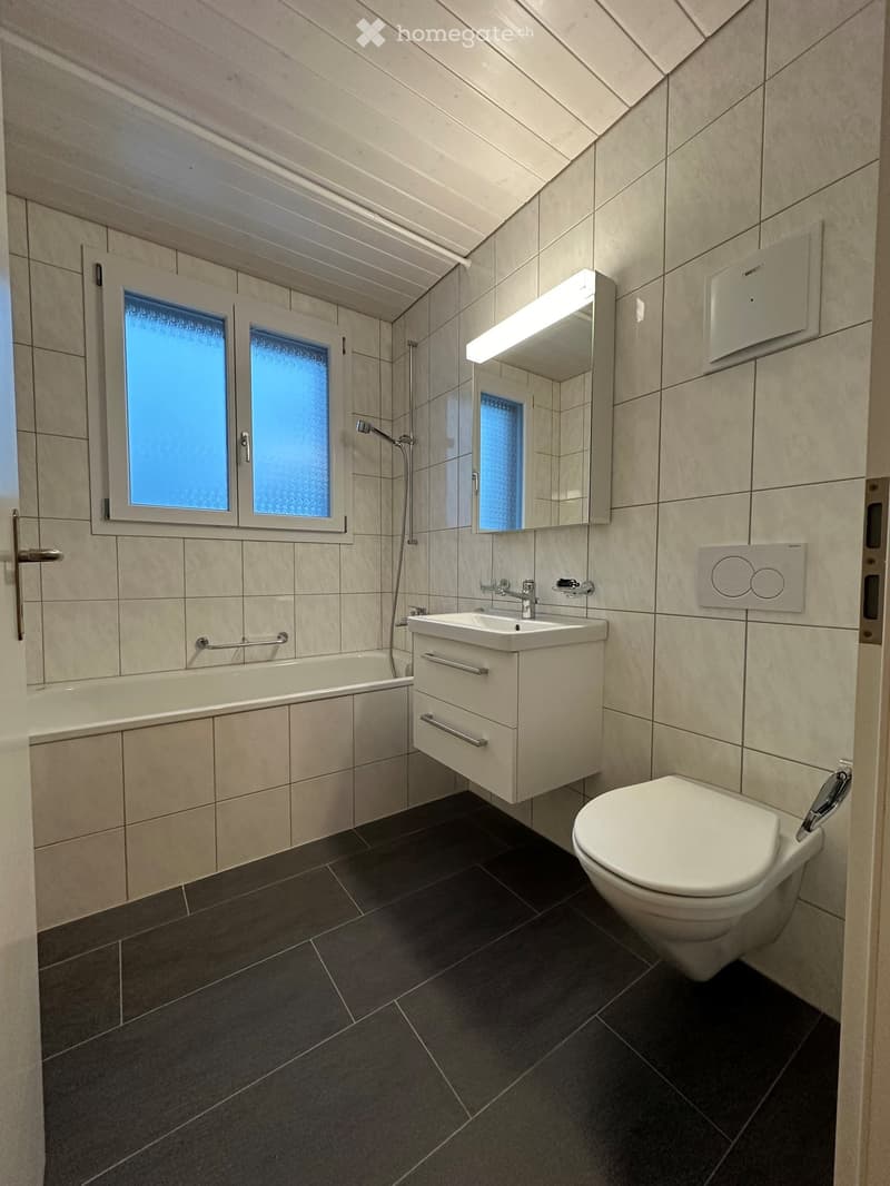 Bathroom with white tiles, bathtub, sink, mirror, and toilet. Shower head and faucet are mounted on the wall.
