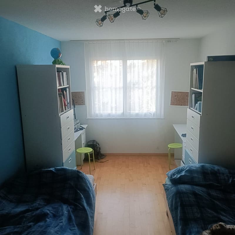 A well-lit bedroom with two beds, a small green stool, a cabinet, and a globe on top of another cabinet. The room features wooden flooring and large windows with white curtains.