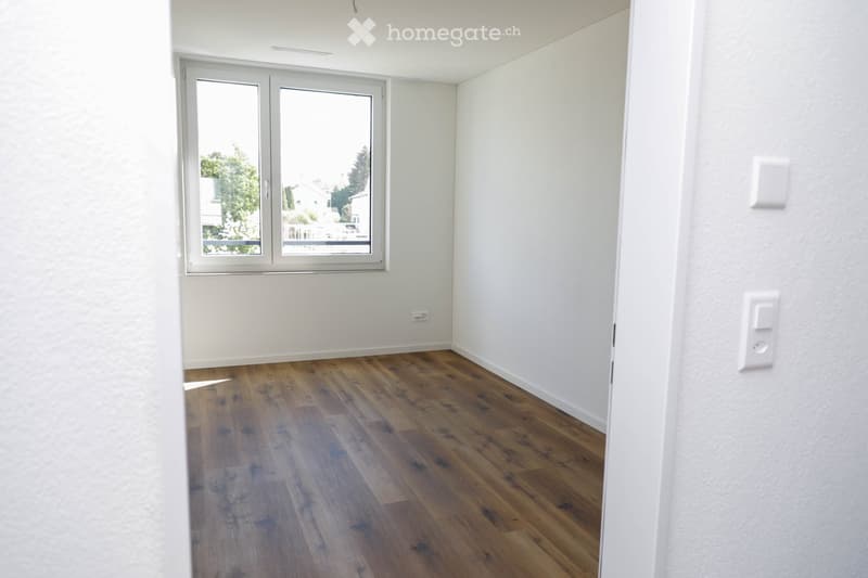 Empty room with light-colored walls, wooden flooring, and a large window with white frames, offering a view of the outside.