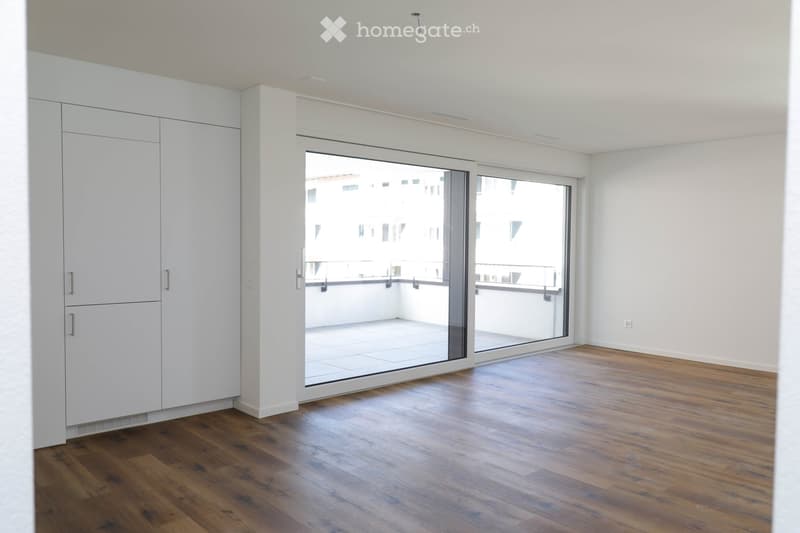 Empty room with wooden flooring, white walls, large sliding glass doors leading to a balcony, and built-in white cabinetry.