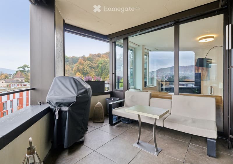 Balcony with a sofa, table, chair, and covered grill, overlooking a scenic view of trees and buildings.