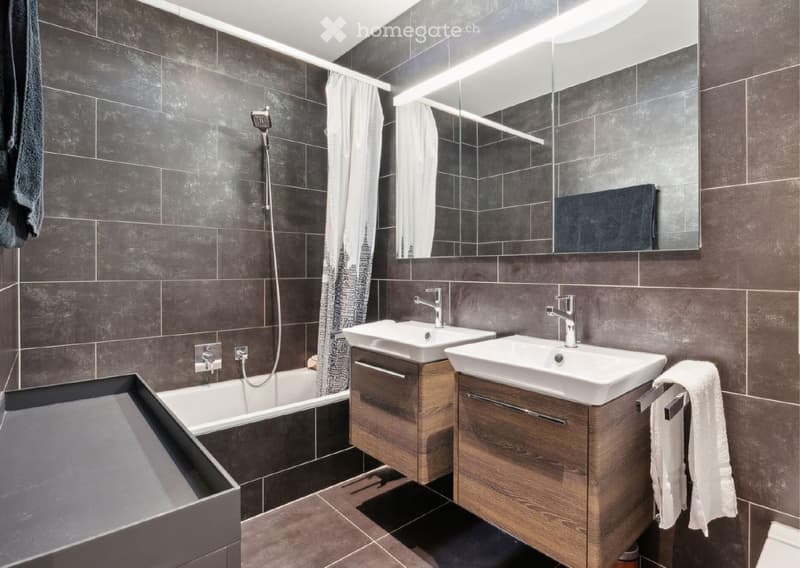 Modern bathroom with dark grey tiles, double sink vanity, bathtub, and large mirror. Includes modern fixtures and towels.
