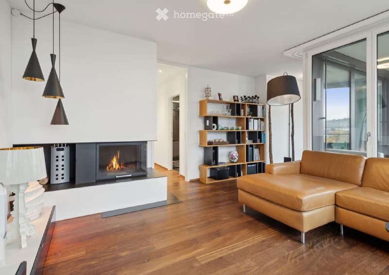 Modern living room with a leather sofa, fireplace, bookshelf, and pendant lights. Large windows offer a scenic view.