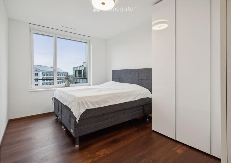 Modern bedroom with a grey upholstered bed, white bedding, wooden floor, large window with city view, and a white wardrobe.