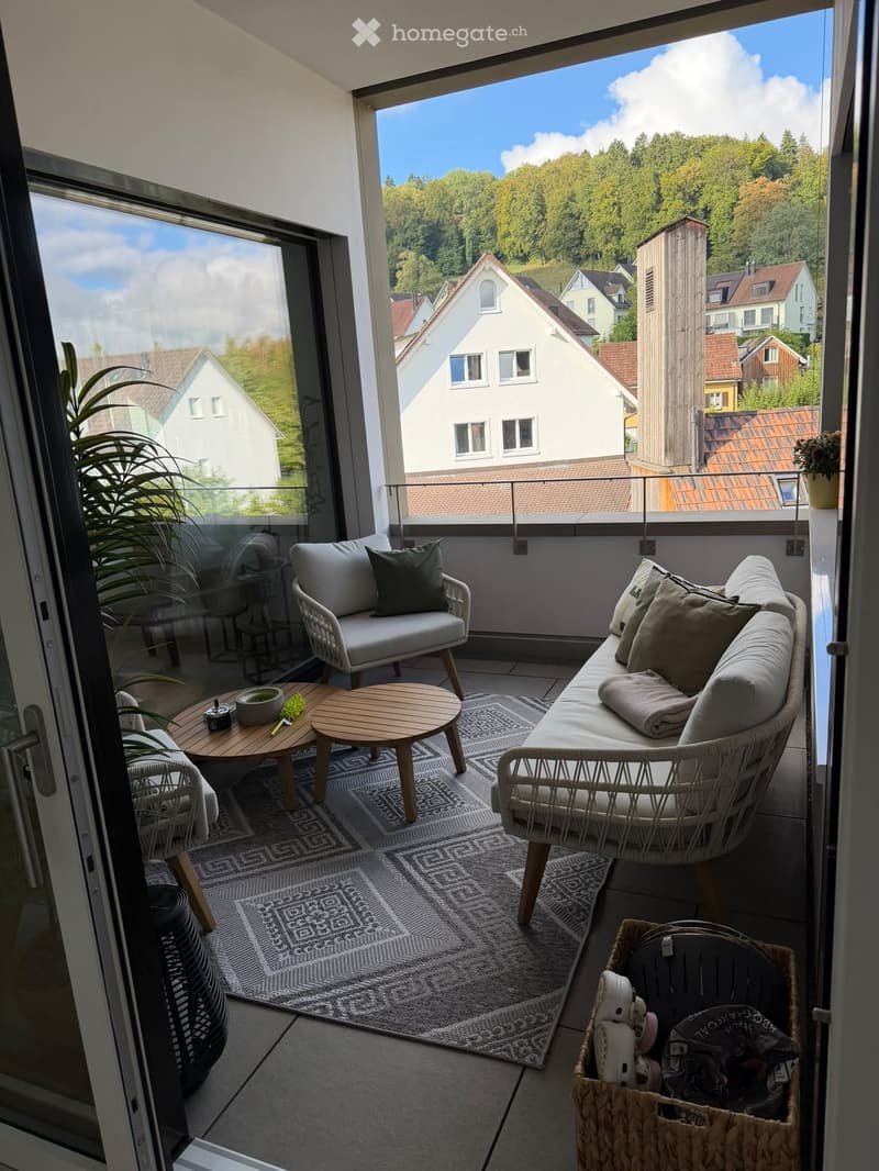 Modern balcony with a view of the town, featuring a sofa, chairs, a small table, a patterned rug, and a woven basket with items.