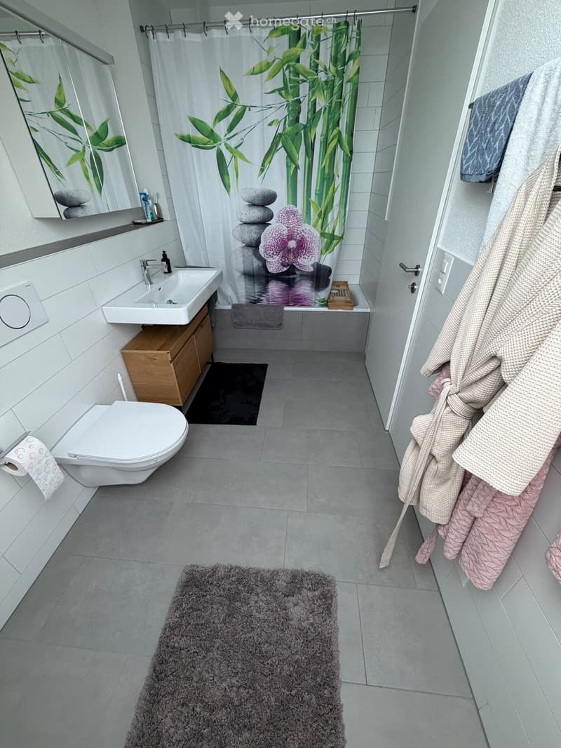 Modern bathroom with white tiles, a sink, toilet, mirror, and a shower area with a bamboo-patterned curtain. A fluffy grey rug is on the floor, and robes and towels are hanging on the door.