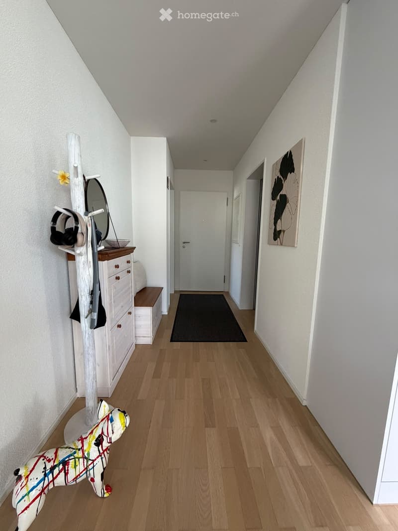 A well-lit hallway with wooden flooring, white walls, and a black runner. Features include a coat rack with a mirror, a small bench, and a colorful abstract sculpture on the floor.