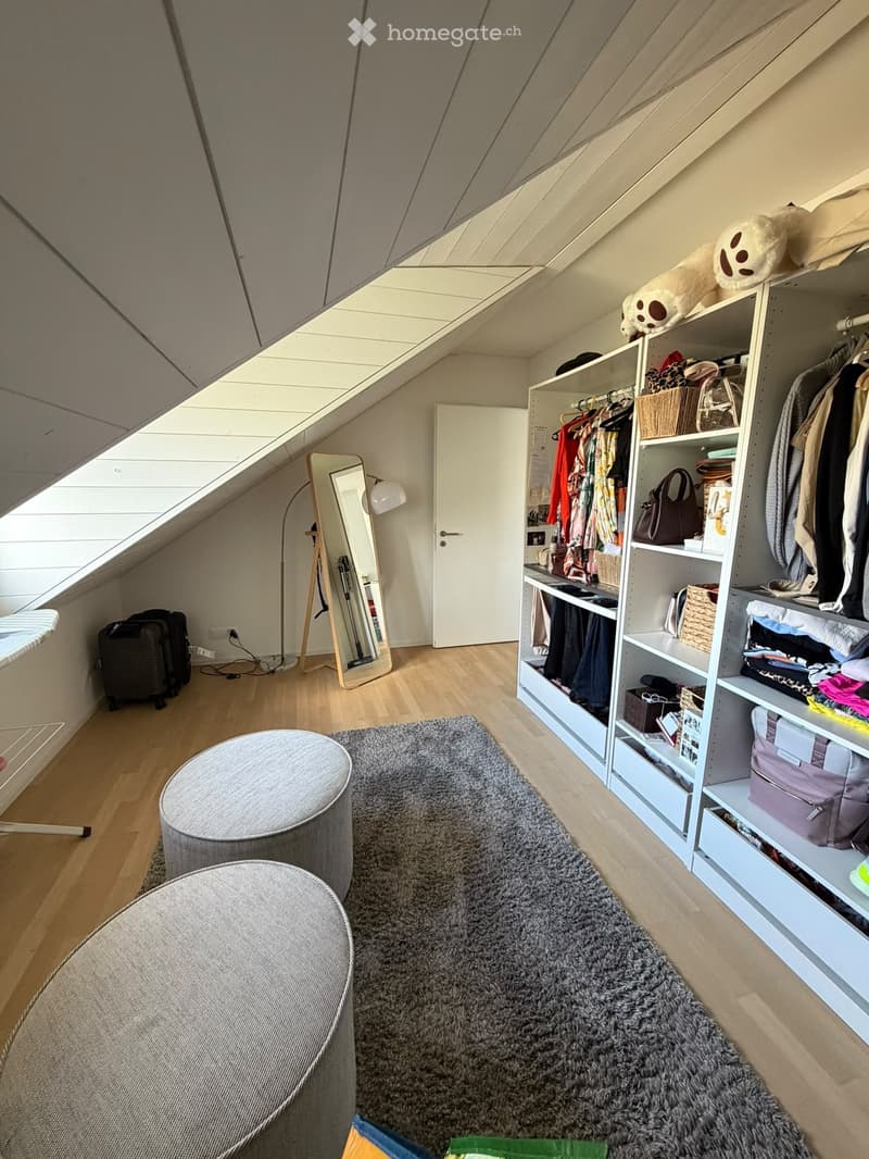 Cozy attic bedroom with wooden floors, grey shaggy rug, two round stools, large mirror, wardrobe with hanging clothes and shelves, and a suitcase near the window.