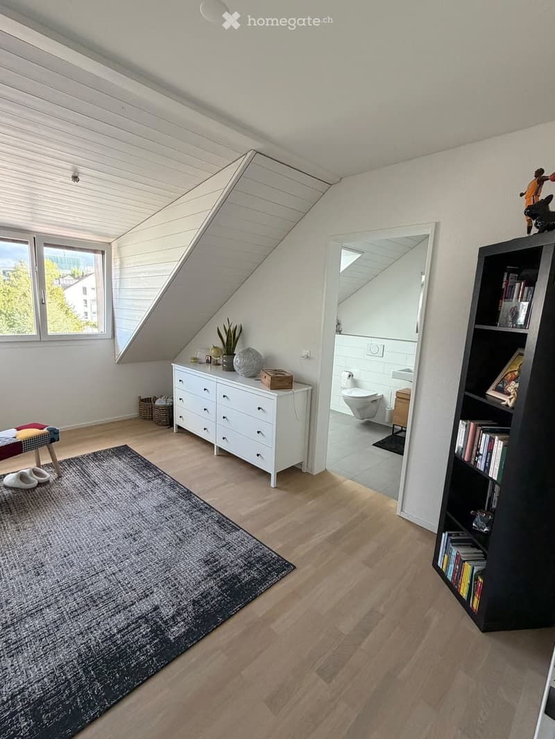 Bright attic room with wooden flooring, slanted ceiling, large window, and a small bathroom visible through an open doorway. Contains a white drawer unit, black bookshelf, grey rug, and decorative items.