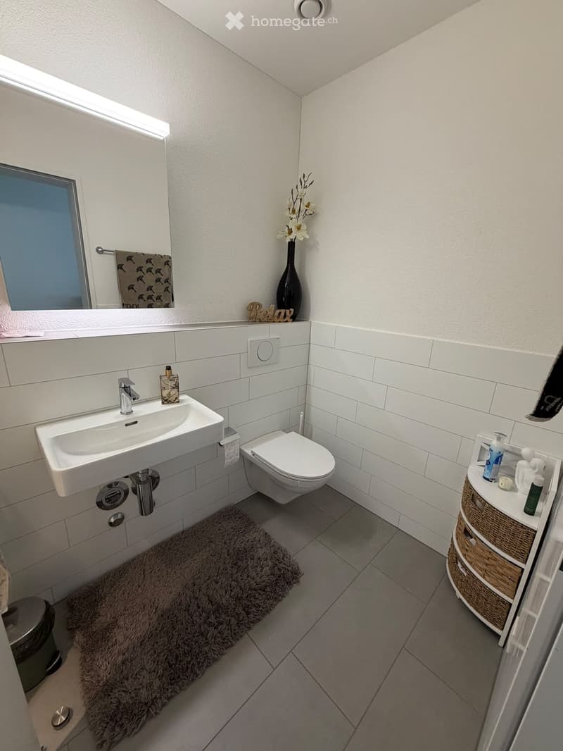 Modern bathroom with white tiles, a sink, toilet, mirror, and a wicker basket holding toiletries. A fluffy rug is on the floor, and a vase with flowers sits on the windowsill.