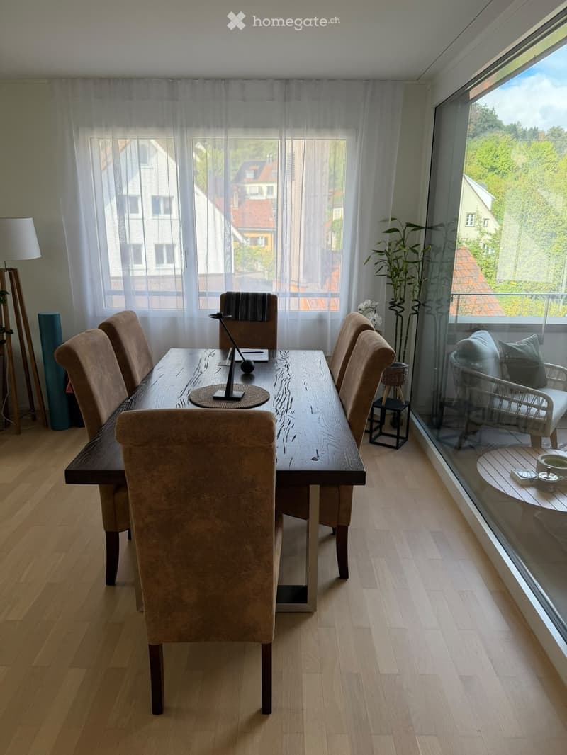 Dining room with a long wooden dining table, six upholstered chairs, a candle holder, a side table, and a small plant near the large window with a view of the neighborhood.