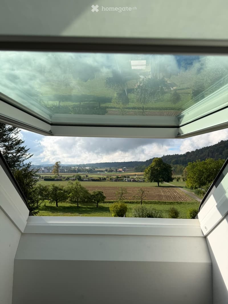 A modern, open skylight or roof terrace with expansive views of a rural landscape, including fields, trees, and distant hills.