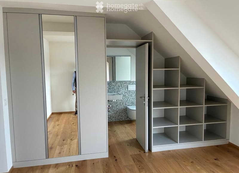 Spacious attic room with wooden flooring, built-in shelving unit, large mirrored closet, and an adjacent modern bathroom with a sink, toilet, and tiled walls.
