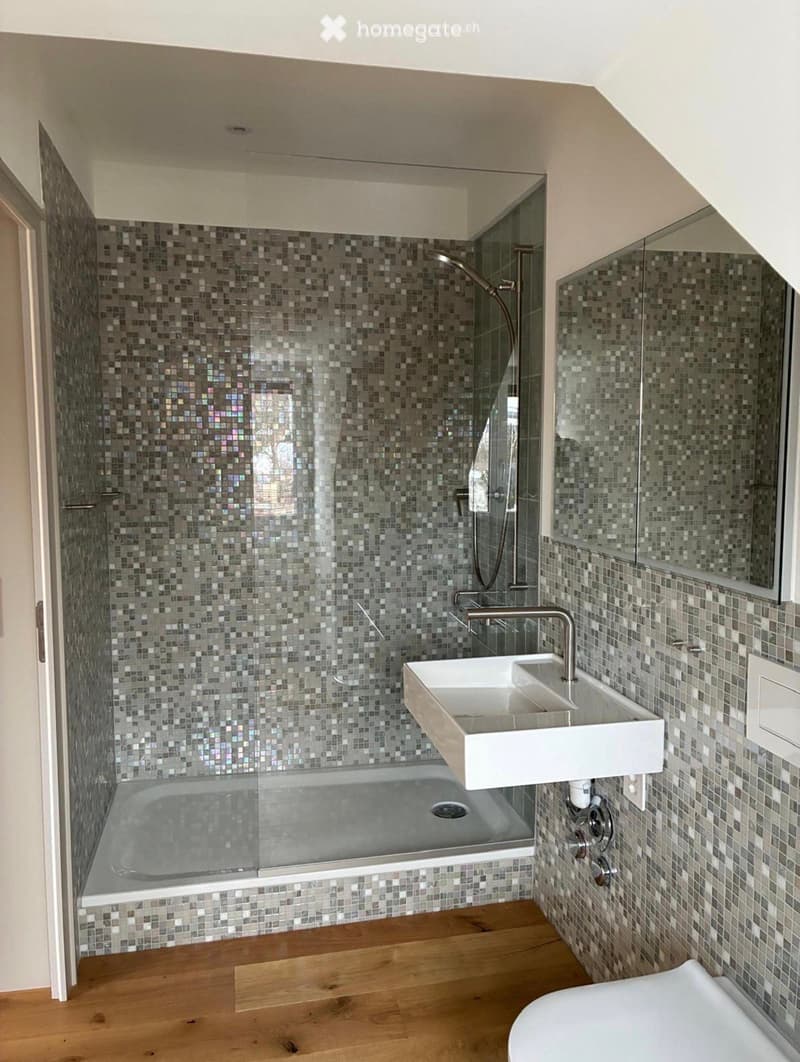 Modern bathroom with mosaic tile walls, a glass-enclosed bathtub, a floating double sink, and a toilet. The space features wooden flooring and a sleek, contemporary design.