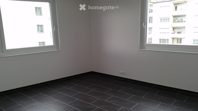 Empty room with dark tiled floor and large windows showing a view of neighboring buildings.