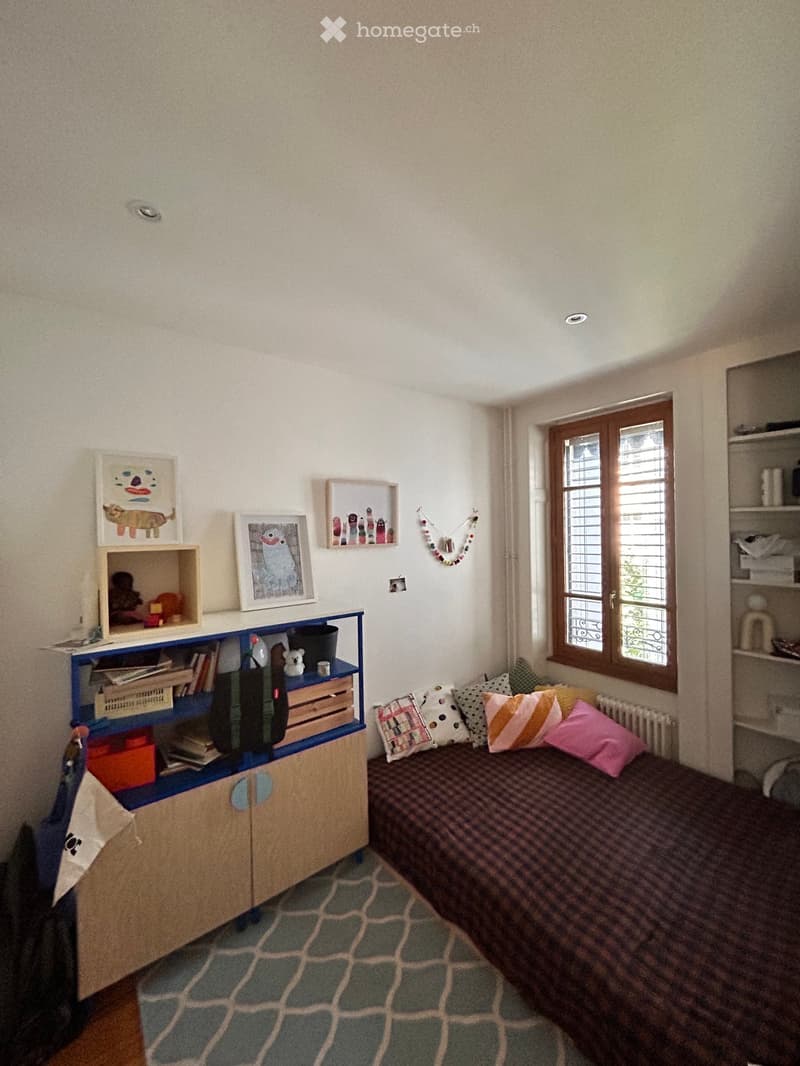 A small, cozy bedroom with a single bed, colorful pillows, a wooden cabinet, shelves, and picture frames on the wall.