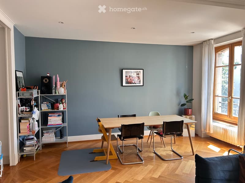 A modern, well-lit room with wooden flooring, a large table surrounded by chairs, a bookshelf filled with books and decorative items, and a cozy seating area with a sofa.