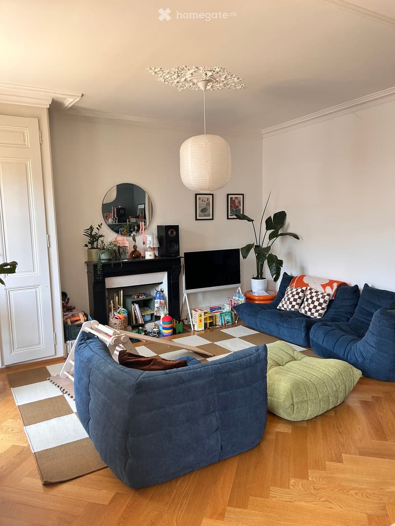 Living room with wooden flooring, a blue sectional sofa, ottoman, coffee table, fireplace, mirror, speaker, television, pendant light, and plants.