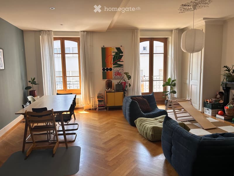 Living room with wooden flooring, large windows, sofa, ottoman, chairs, table, potted plants, and decorative items.