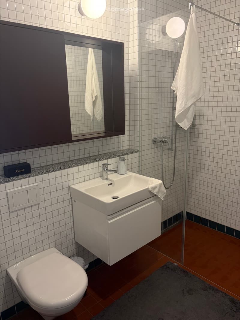 Modern bathroom with white tiles, a sink with a modern faucet, a toilet, a large mirror, a glass-enclosed shower, and a towel hanging on a rod.