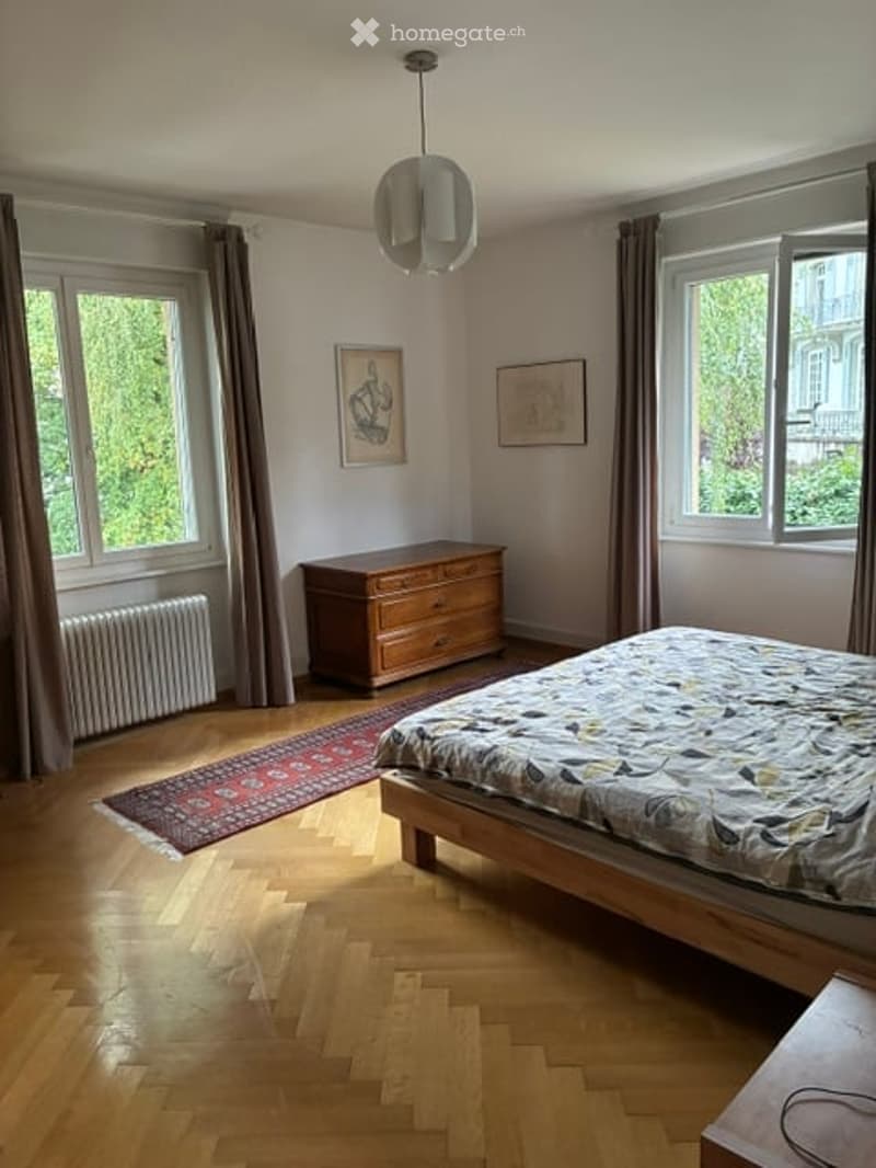 Bright bedroom with wooden flooring, large windows with curtains, a bed with patterned bedding, a wooden chest of drawers, a small rug, and two picture frames on the wall.
