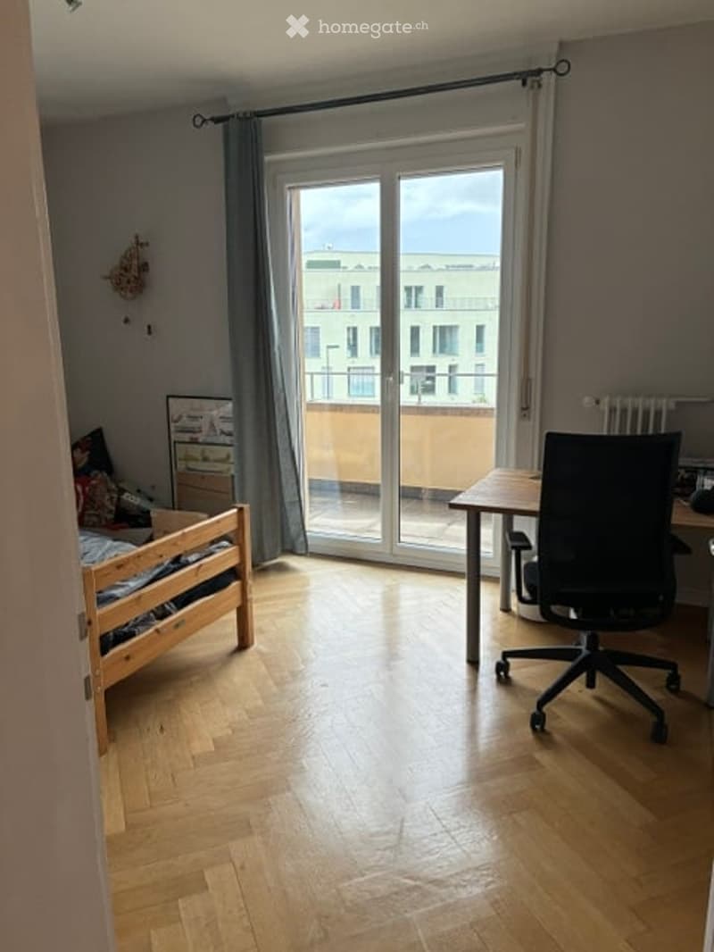 Bright bedroom with wooden flooring, large sliding glass door leading to a balcony, bed, desk, and chair.