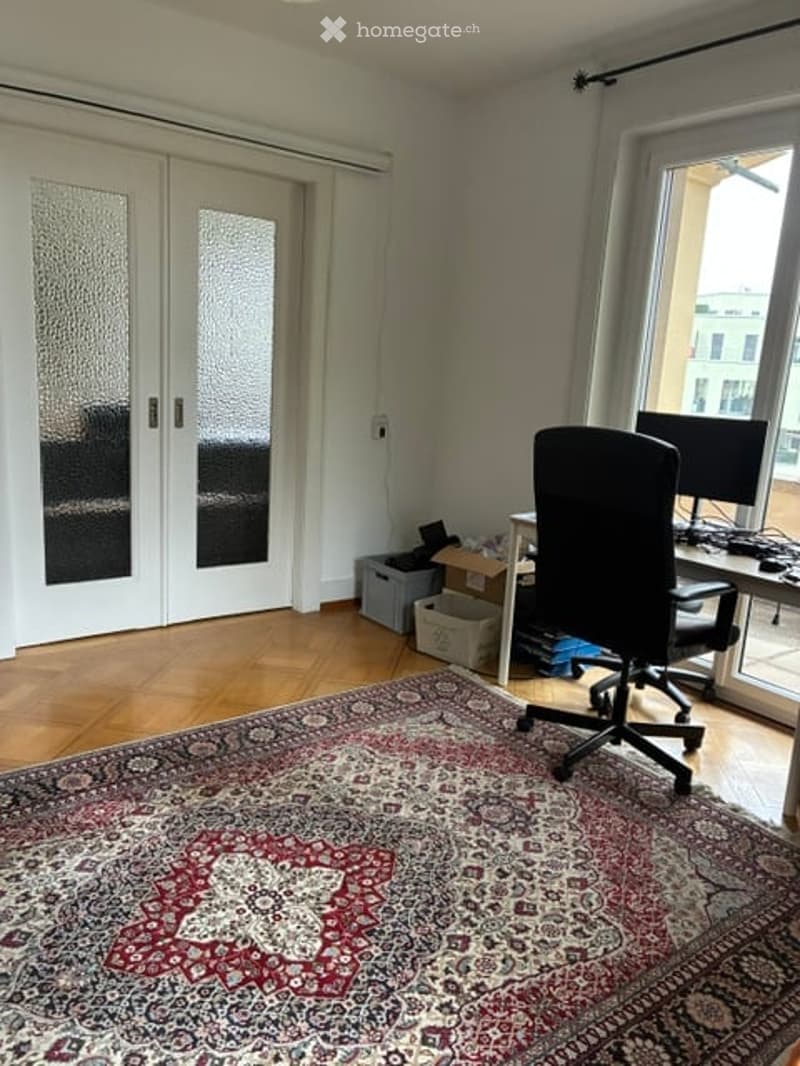 Office space with a large window offering a city view, a patterned rug, a black office chair, a desk with a computer, and storage boxes.
