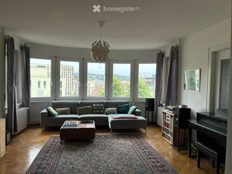 Bright living room with large windows offering a city view, featuring a grey sectional sofa, coffee table, piano, and wooden flooring.