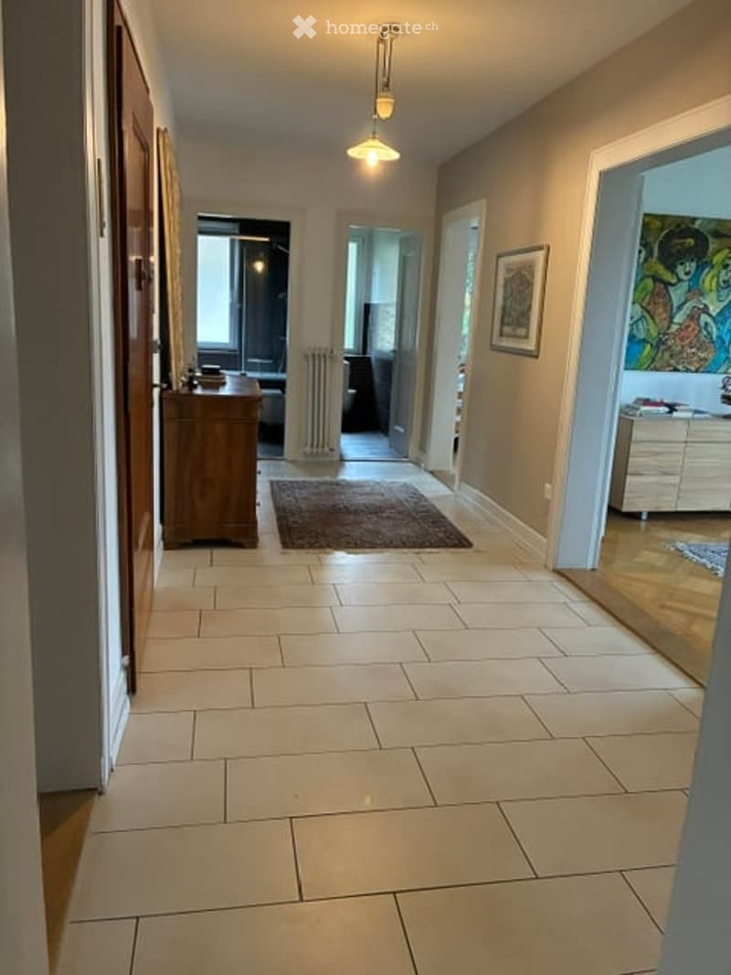A well-lit hallway with tiled flooring, beige walls, and several doors leading to different rooms. There are small rugs and a few pieces of furniture visible in the background.