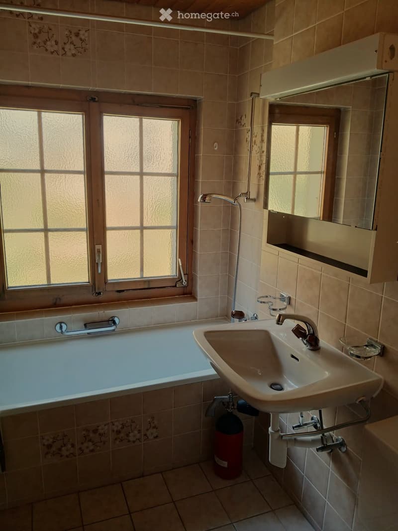 Bathroom with beige tiled walls and floor, featuring a bathtub, sink with faucet, mirror cabinet, and a fire extinguisher on the floor.