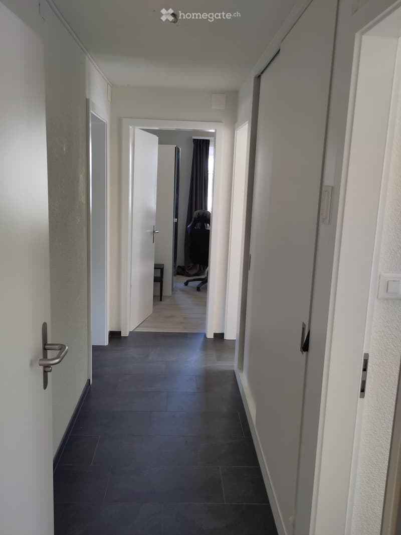 A long, narrow hallway with dark tiled flooring, white walls, and several doors on both sides. The hallway leads to an open doorway at the end, revealing a glimpse of another room with a chair and a curtain.