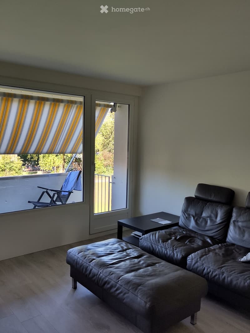 Modern living room with a black leather sofa, coffee table, and a balcony with a blue folding chair.