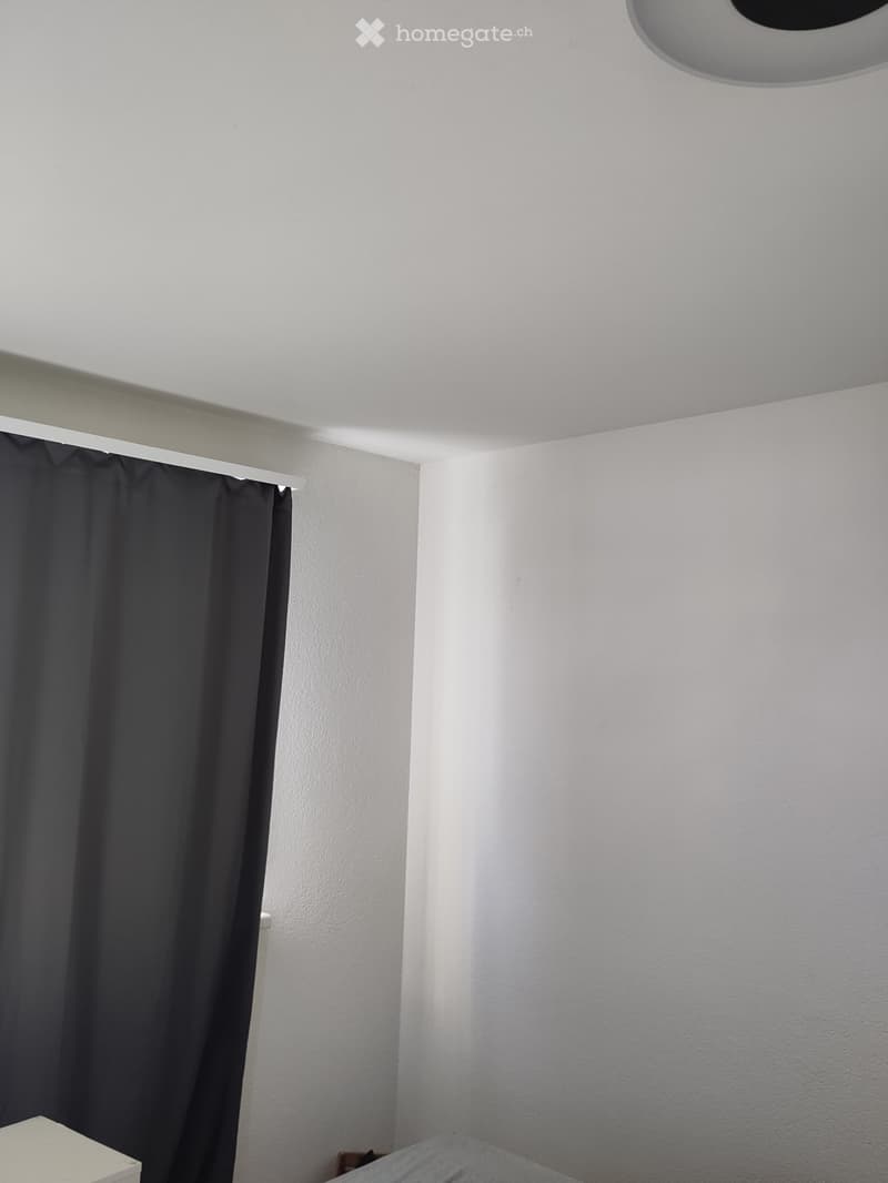 Completely empty room with bare white walls, a dark curtain, and a white ceiling light fixture.