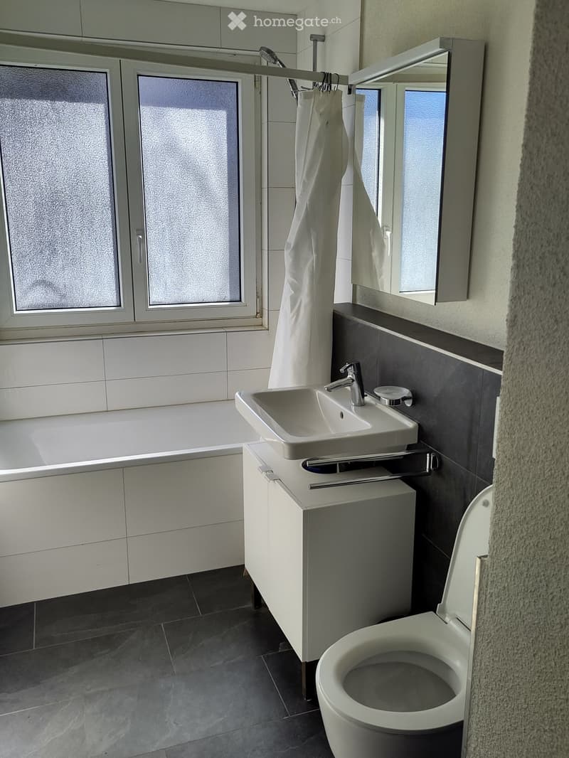 Modern bathroom with a bathtub, sink, toilet, and mirror. Features white tiles, dark grey lower wall tiles, and a large window with frosted glass.