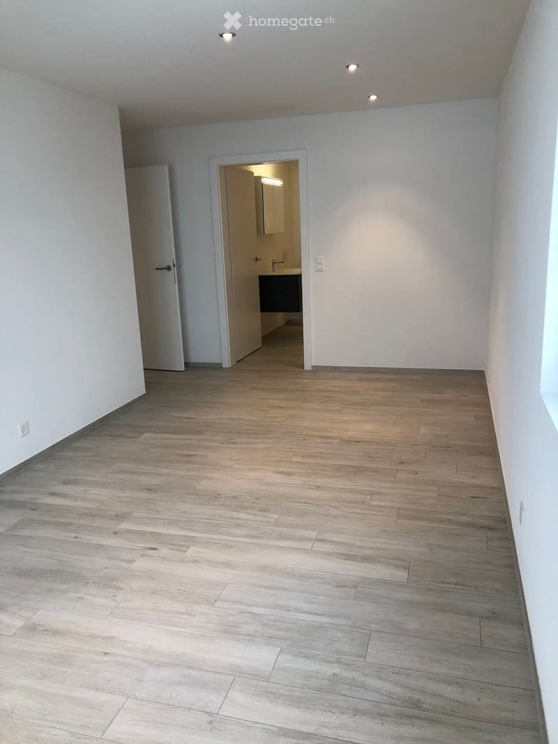 Empty room with light wooden flooring and white walls, featuring a partially open door leading to a small bathroom with a sink and mirror.