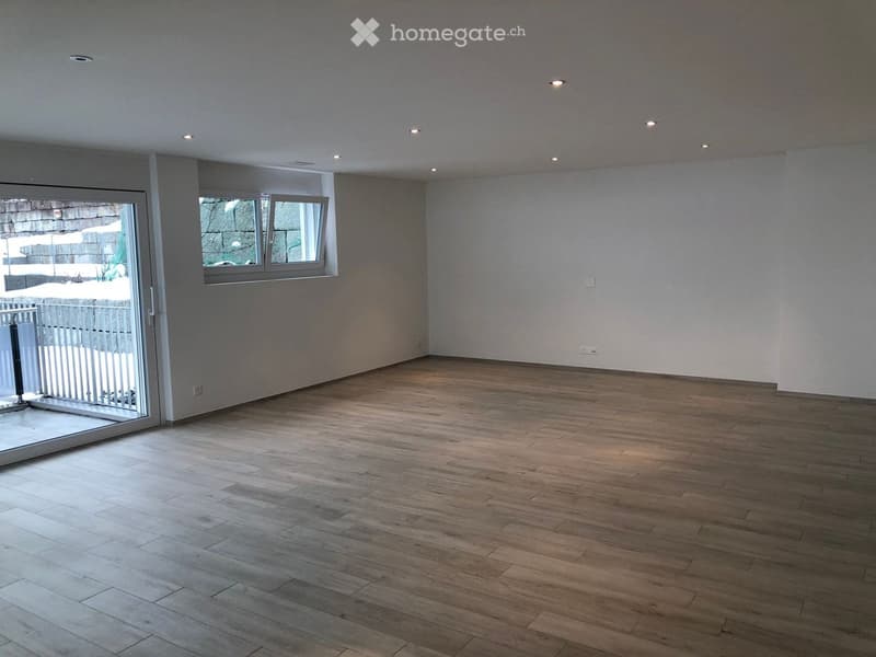 Empty room with wooden flooring, white walls, and recessed ceiling lights. Features a large window and a sliding glass door leading to an outdoor area.