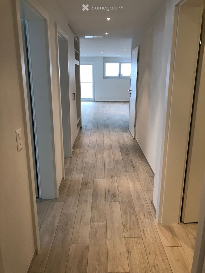 Empty hallway with wooden flooring, white walls, and recessed lighting. Doors lead to adjacent rooms, and a window at the end provides natural light.