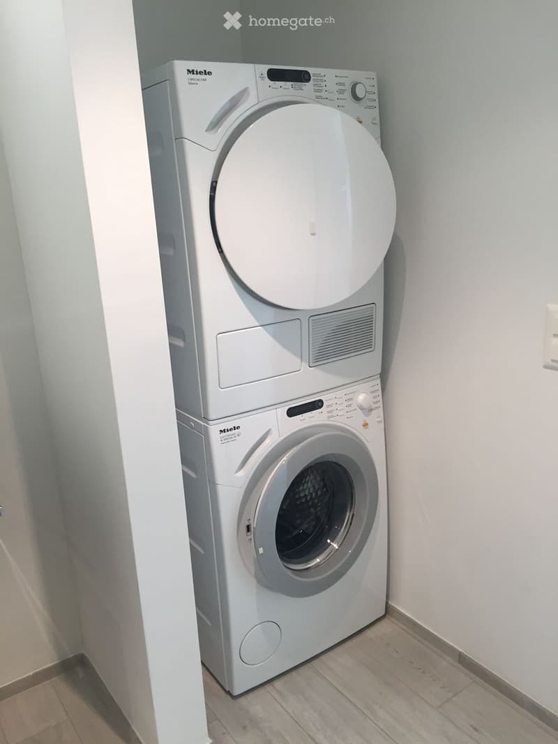 A compact bathroom with a white washing machine and dryer stacked vertically, featuring a control panel and a circular drum. The floor is tiled, and the walls are painted light gray.