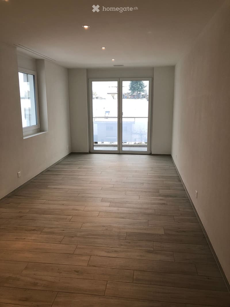 Empty hallway with wooden flooring, white walls, and recessed lighting. Features a large sliding glass door leading to a balcony area.