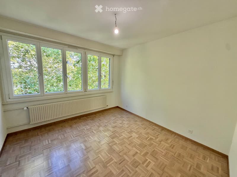 Empty room with wooden herringbone flooring, large windows with white frames, white walls, and a single ceiling light fixture.