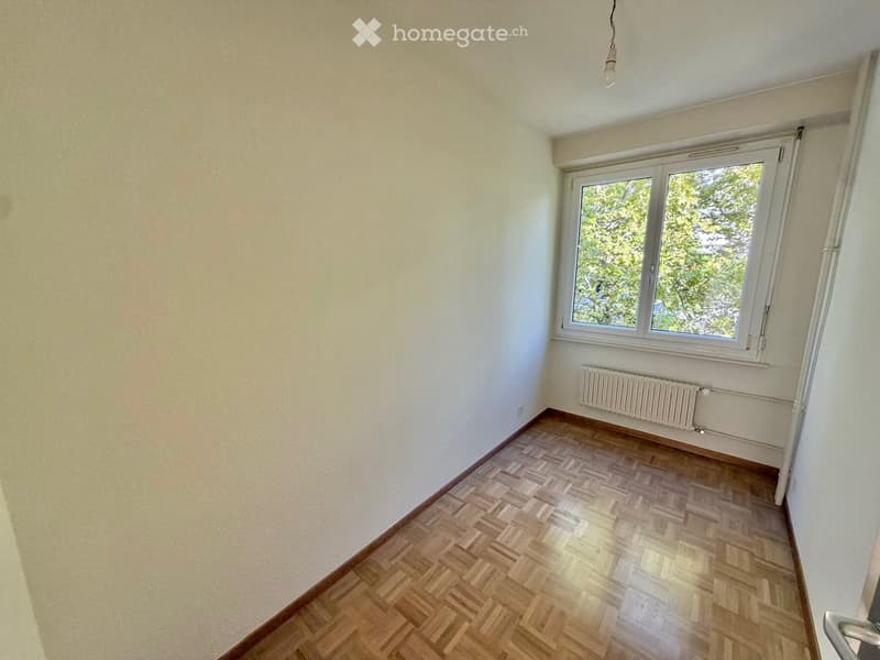 Empty room with wooden herringbone flooring, white walls, a window with white frame, and a radiator below the window.