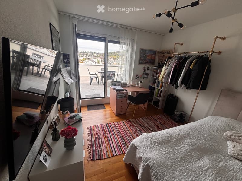 Cozy bedroom with a bed, desk, chair, mirror, bookshelf, clothing rack, and a view of a balcony area with outdoor furniture.