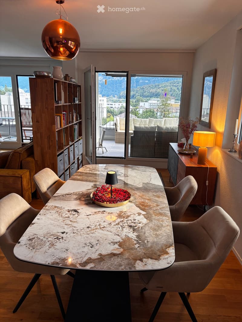 Modern living room with a large marble dining table, chairs, a sofa, bookshelf, and a view of the mountains through large windows.