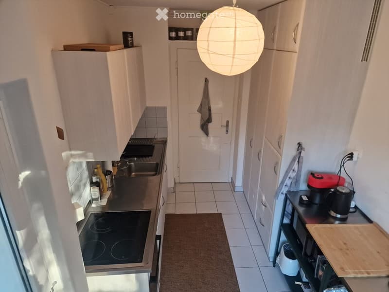 Compact kitchen with white cabinets, induction stove, microwave, coffee maker, toaster, and various small appliances. Features a hanging paper lantern light fixture and a small wooden cutting board on the counter.